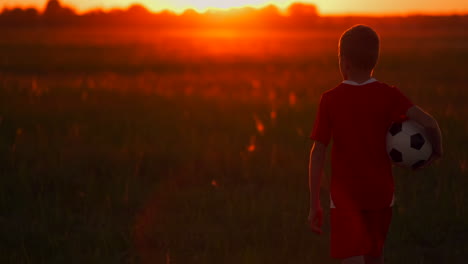 The-boy-is-on-the-field-with-the-ball-in-his-hands-looking-at-the-sunset-and-dreaming-of-a-football-career.-the-camera-follows-the-boy.
